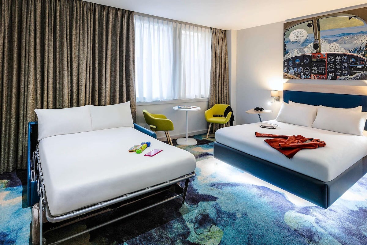 A hotel room with two beds, a desk, a chair, a small table, and a window with curtains.