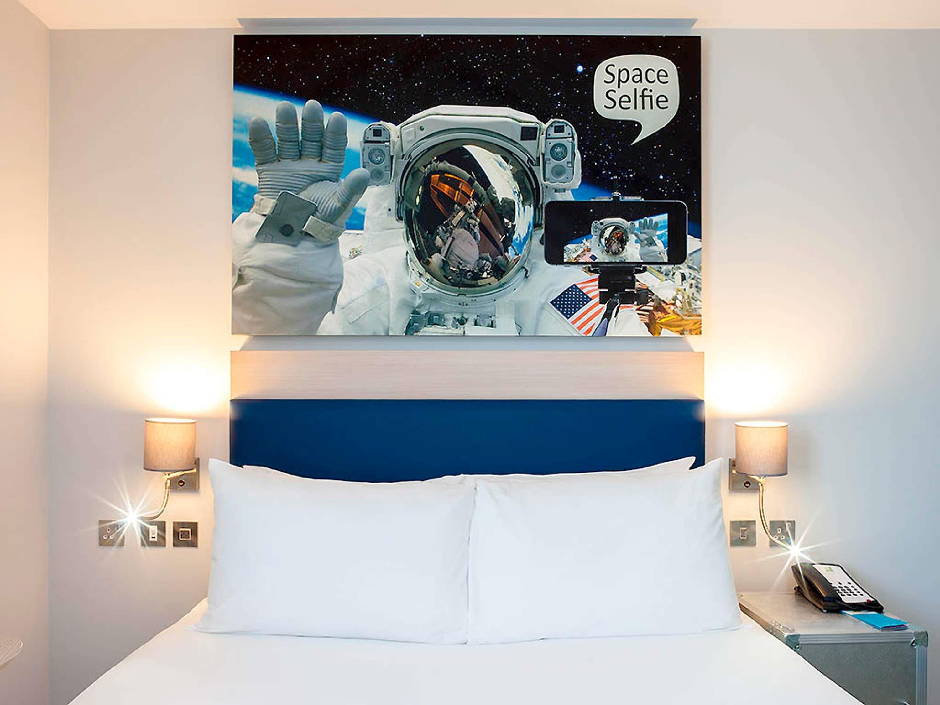 A bedroom with a large bed, two bedside lamps, and a wall-mounted painting of an astronaut taking a selfie in space.