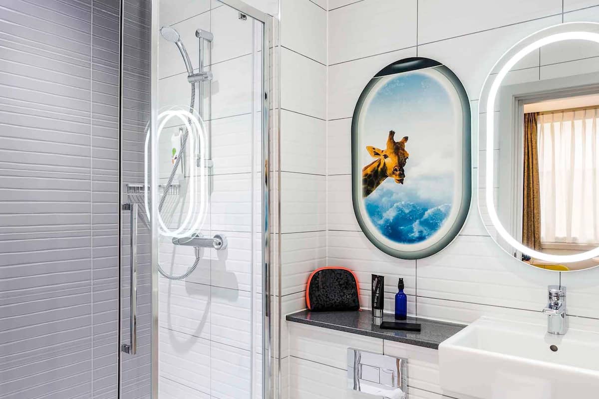 A bathroom with a glass shower enclosure, a round mirror with a giraffe image, and a white sink with a towel rack.