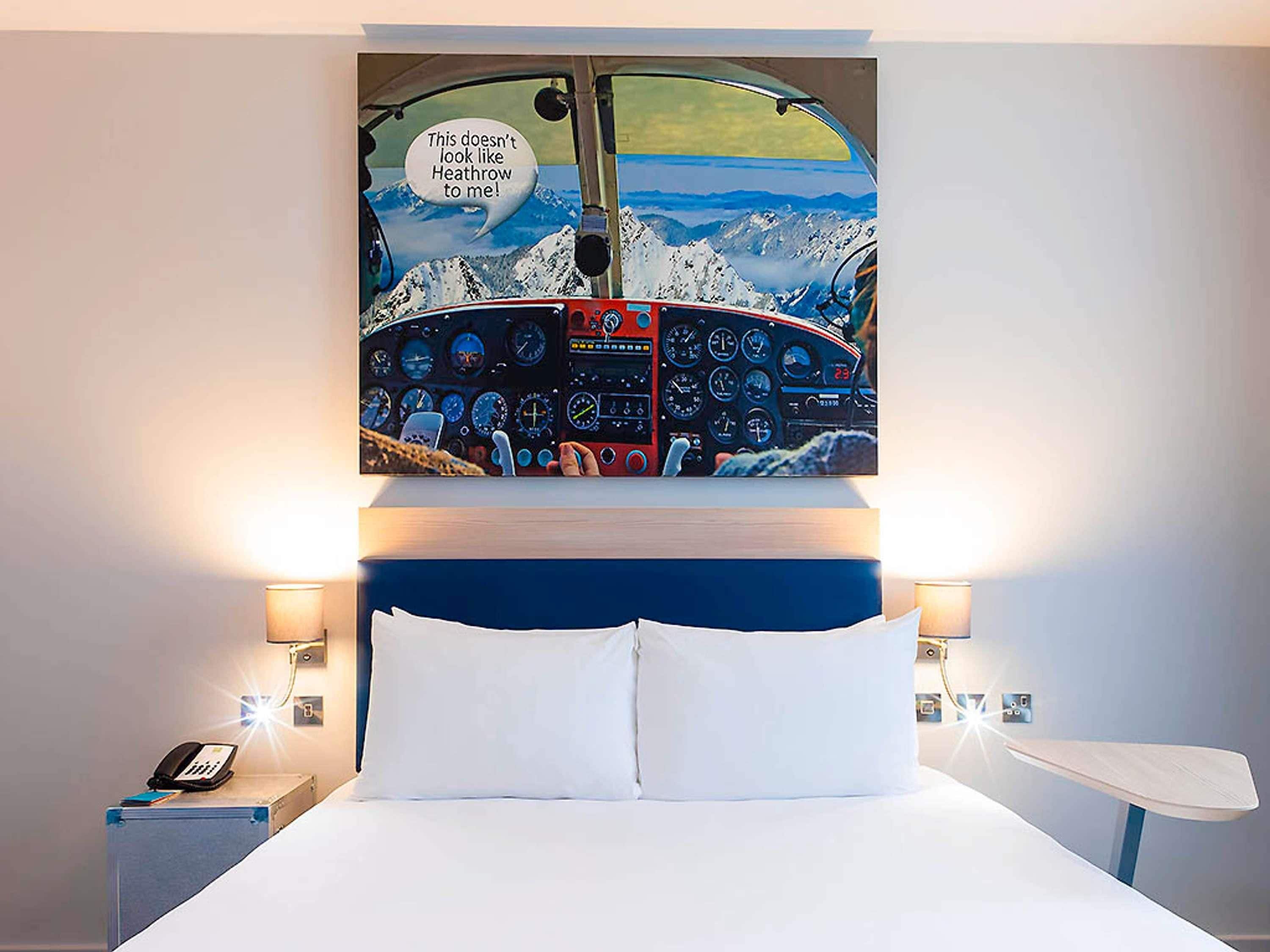 A hotel room with a bed, a headboard with a painting of an airplane cockpit, bedside lamps, and a nightstand with a phone.