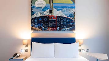 A hotel room with a bed, a headboard with a painting of an airplane cockpit, bedside lamps, and a nightstand with a phone.