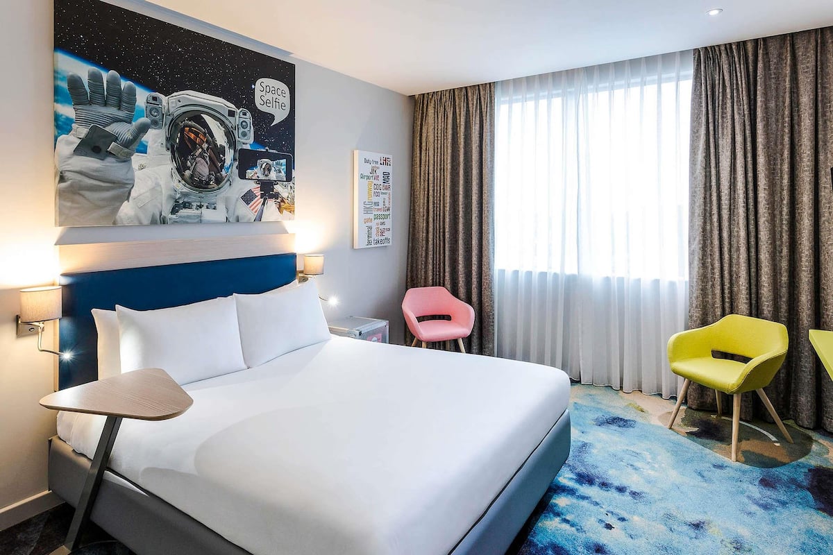 A hotel room with a bed, a wall-mounted space-themed artwork, a window with curtains, and two chairs.