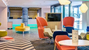 A modern lounge area with colorful seating, a large flat-screen TV, and a patterned floor. - Ibis Styles London Heathrow (West Drayton)