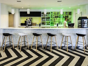 A modern bar area with a bar counter, stools, and a green-lit shelving unit displaying bottles. - Ibis Styles London Heathrow (West Drayton)