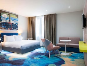 Moderni hotellihuone, jossa on suuri sänky, työpöytä, sohva ja värikäs, abstrakti kuvioitu matto.