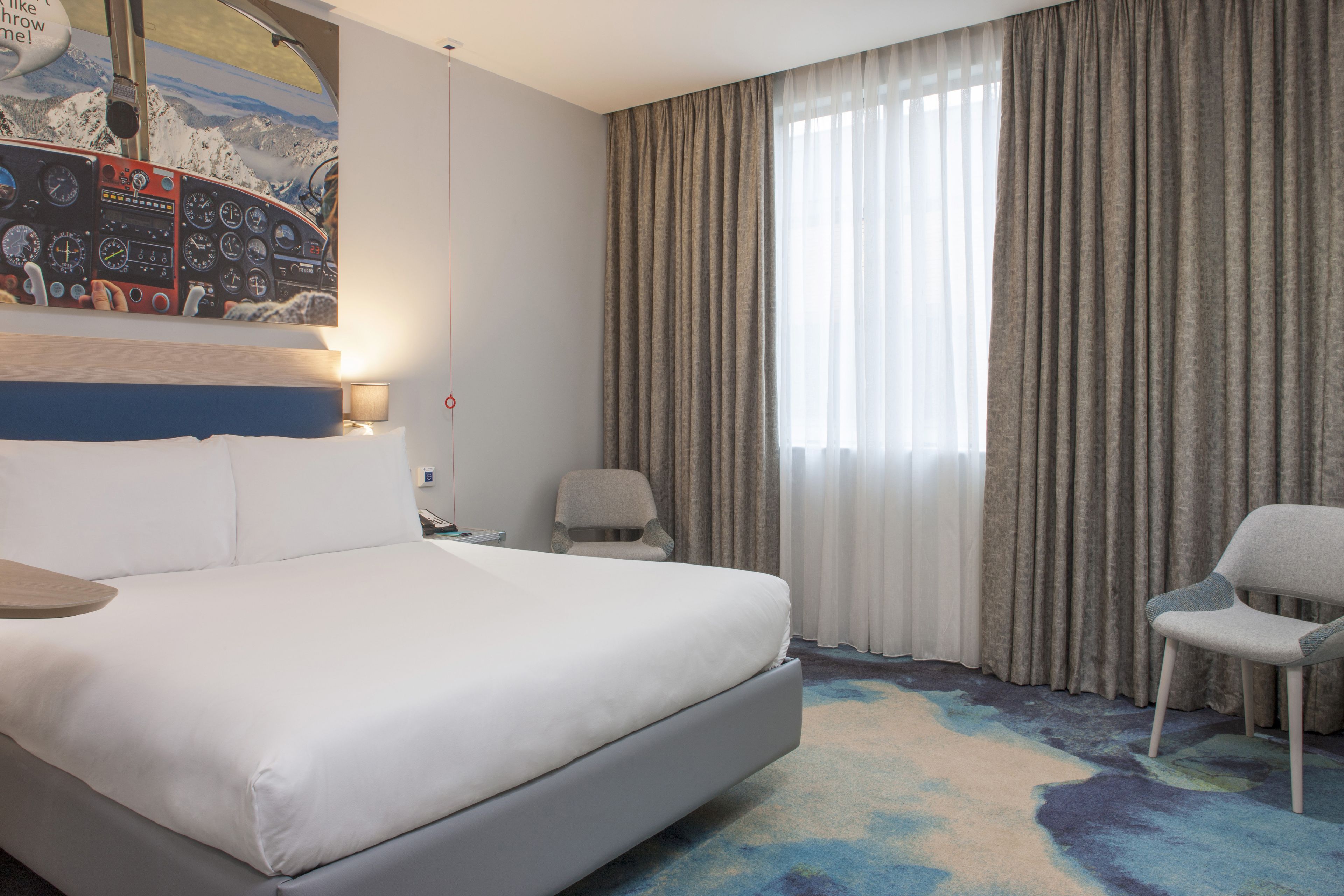 A hotel room with a large bed, a chair, a desk, and a window with curtains.