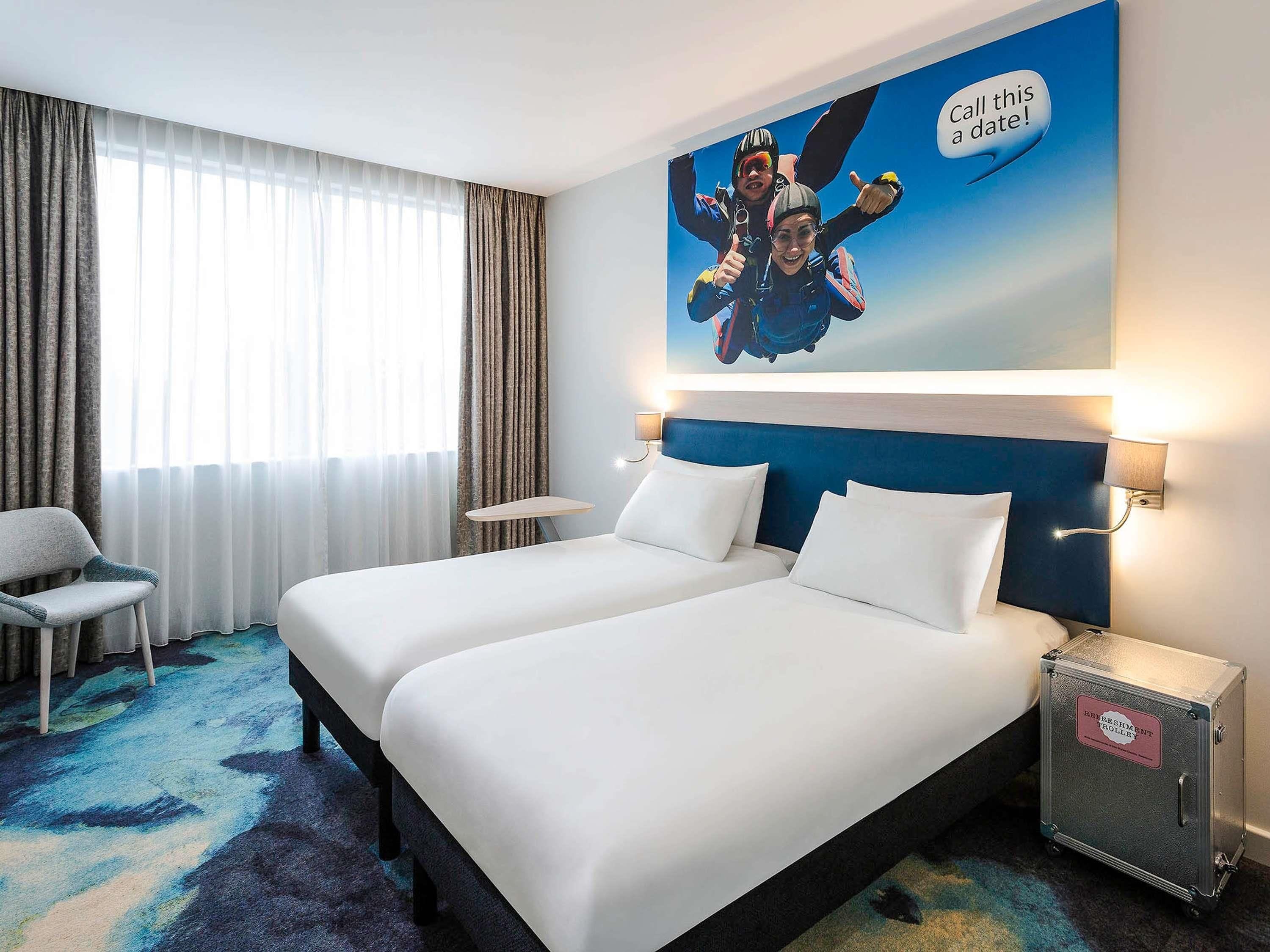 Une chambre d’hôtel avec deux lits, une petite table, une chaise, une table de chevet et une lampe fixée au mur.