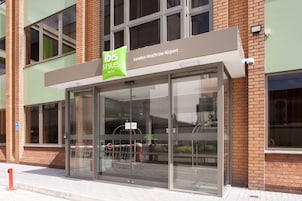 The exterior of the ibis Styles Hotel at London Heathrow Airport, featuring a modern glass facade, a prominent green and white sign, and a clear view of the airport entrance.