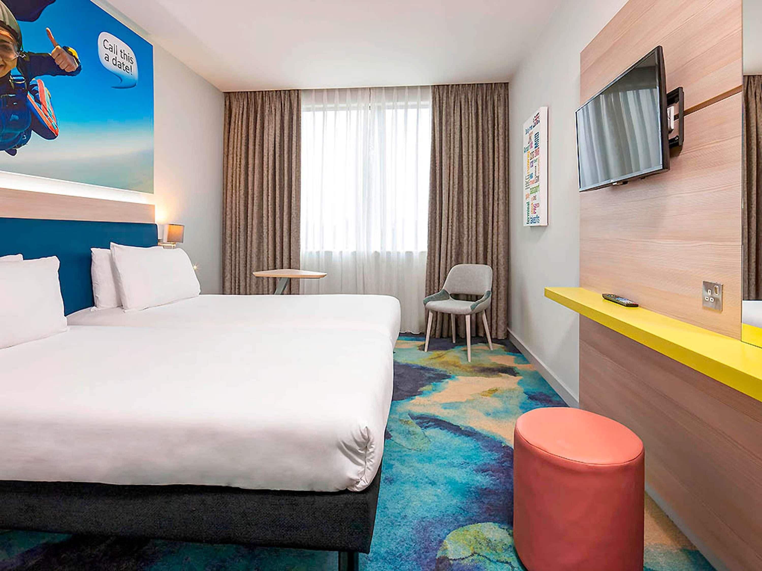 A hotel room with a bed, a desk, a chair, a TV, and a colorful carpet.