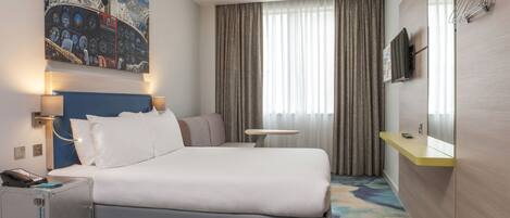 A modern hotel room with a large bed, a desk, a chair, a small table, a TV, and a painting on the wall.