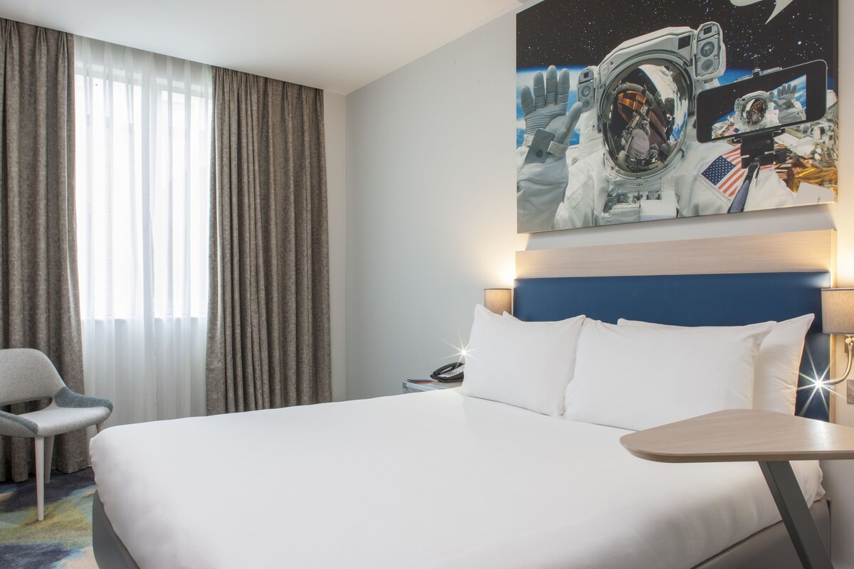 Hotellihuone, jossa on suuri sänky, työpöytä, tuoli ja seinällä kuva astronautista.