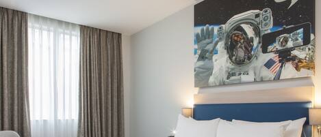 Ein Hotelzimmer mit einem großen Bett, einem Schreibtisch, einem Stuhl und einem Gemälde eines Astronauten an der Wand.