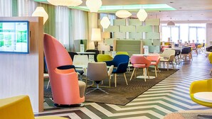A modern hotel lobby with colorful chairs, a large screen displaying a menu, and a patterned floor. - Ibis Styles London Heathrow (West Drayton)