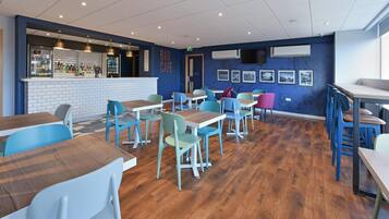 A modern bar and seating area with wooden tables and colorful chairs, a bar counter with a white tiled backsplash, and framed pictures on the walls.