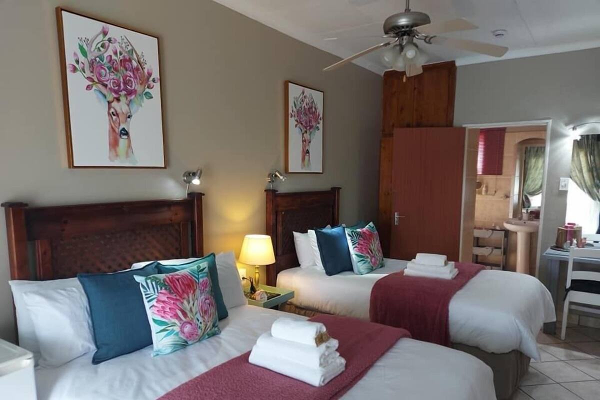 A hotel room with three beds, each with white linens and decorative pillows. There is a ceiling fan, a bedside table with a lamp, and artwork on the walls.