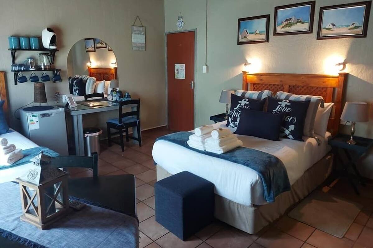 A hotel room with a bed, a desk, a chair, a small table, and a lamp. There are pictures on the wall and a door in the background.