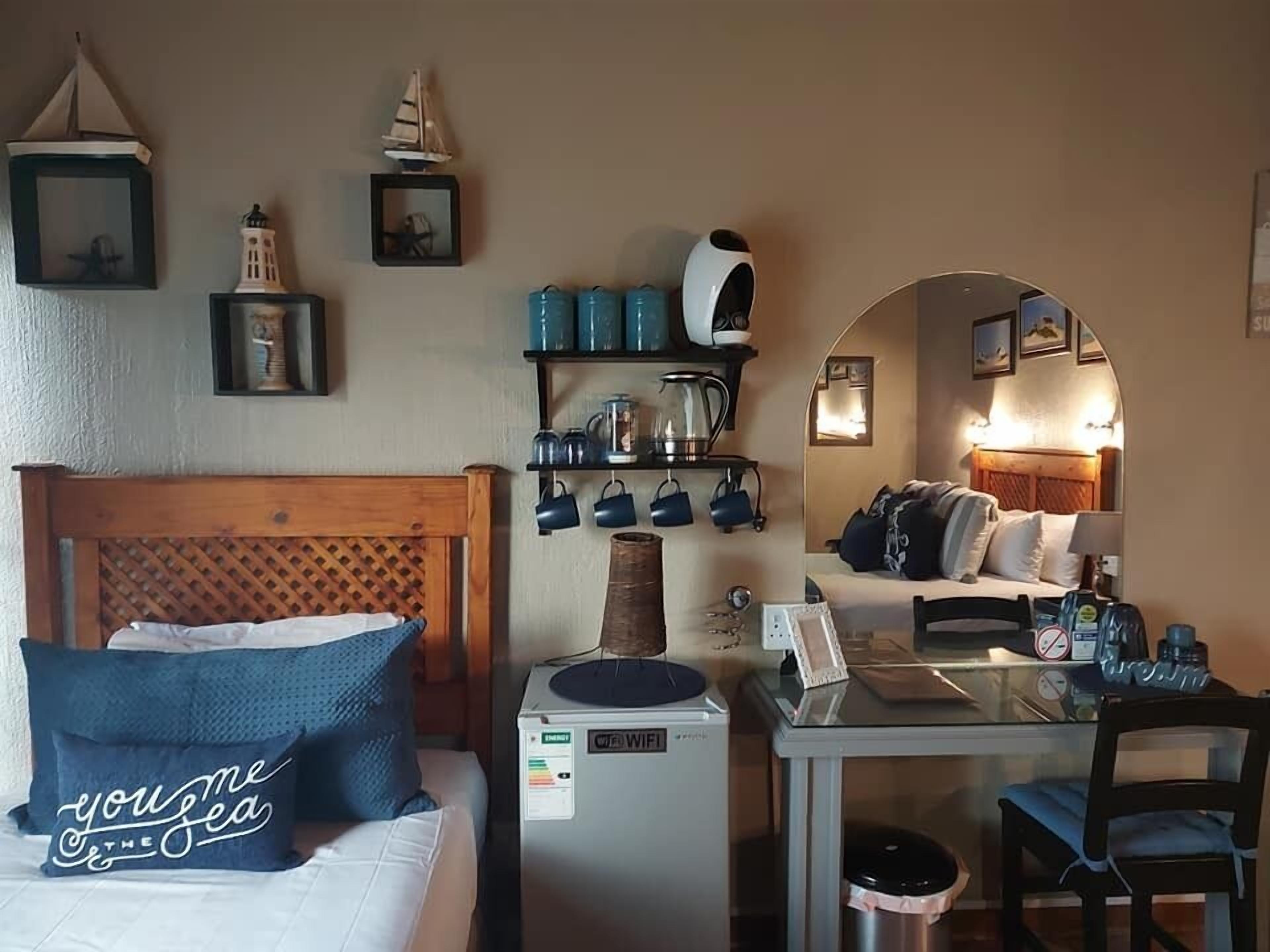 Quarto de hotel com cama, escrivaninha com computador, cafeteira e pequeno aquecedor. Há prateleiras com canecas e uma luminária.