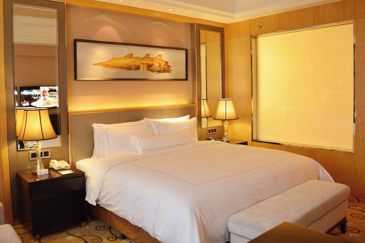 Ein Hotelzimmer mit einem großen Bett, einem Nachttisch mit Lampe, einem Fernseher und einem Gemälde an der Wand.