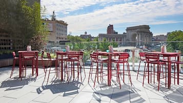 A rooftop terrace with red metal tables and chairs, offering a view of a cityscape including a large arch structure.