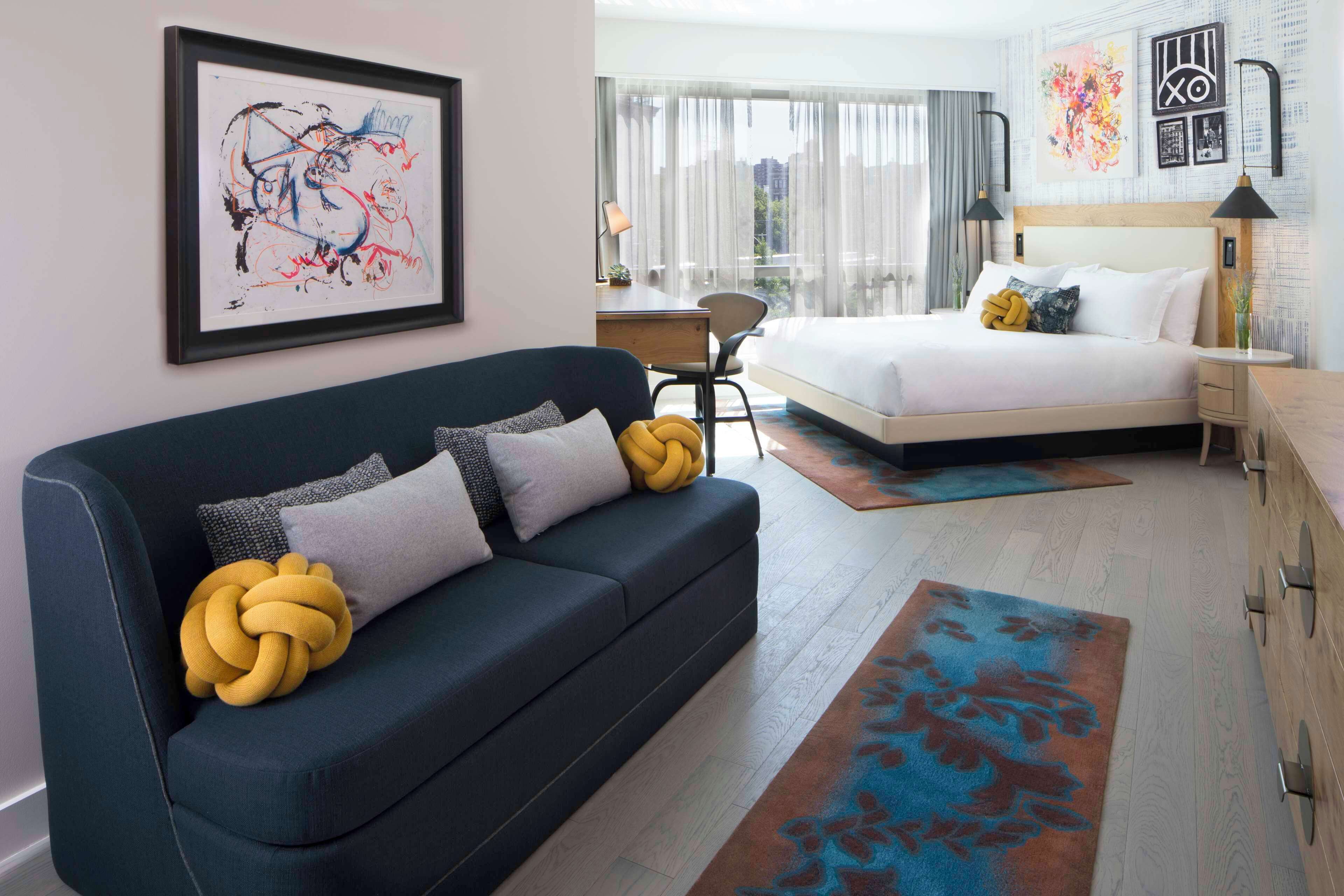 A modern hotel room with a dark blue sofa, a bed with white linens, a large window with curtains, and artwork on the walls.