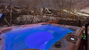 Un bain à remous doté d’un panneau de commande, entouré d’une terrasse offrant une vue sur des collines enneigées au crépuscule.
