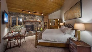 A bedroom with a stone fireplace, a bed, a desk with a chair, and a television. - Trails Edge Lodge (Steamboat Springs)