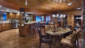A spacious kitchen with wooden cabinetry and a central island, a dining area with leather chairs, and a bar area with a high ceiling and wooden beams. - Trails Edge Lodge (Steamboat Springs)