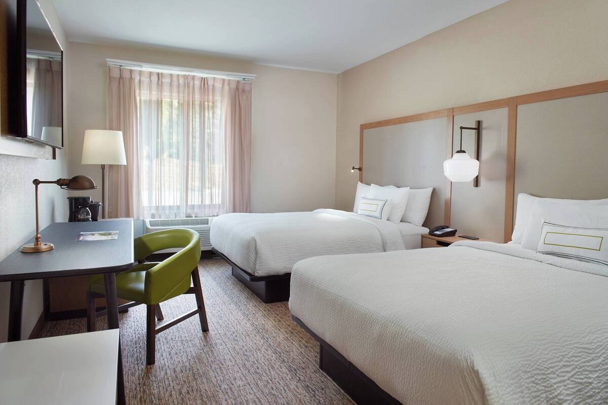Une chambre d’hôtel avec deux lits, un bureau, une chaise, une lampe et une fenêtre avec des rideaux.