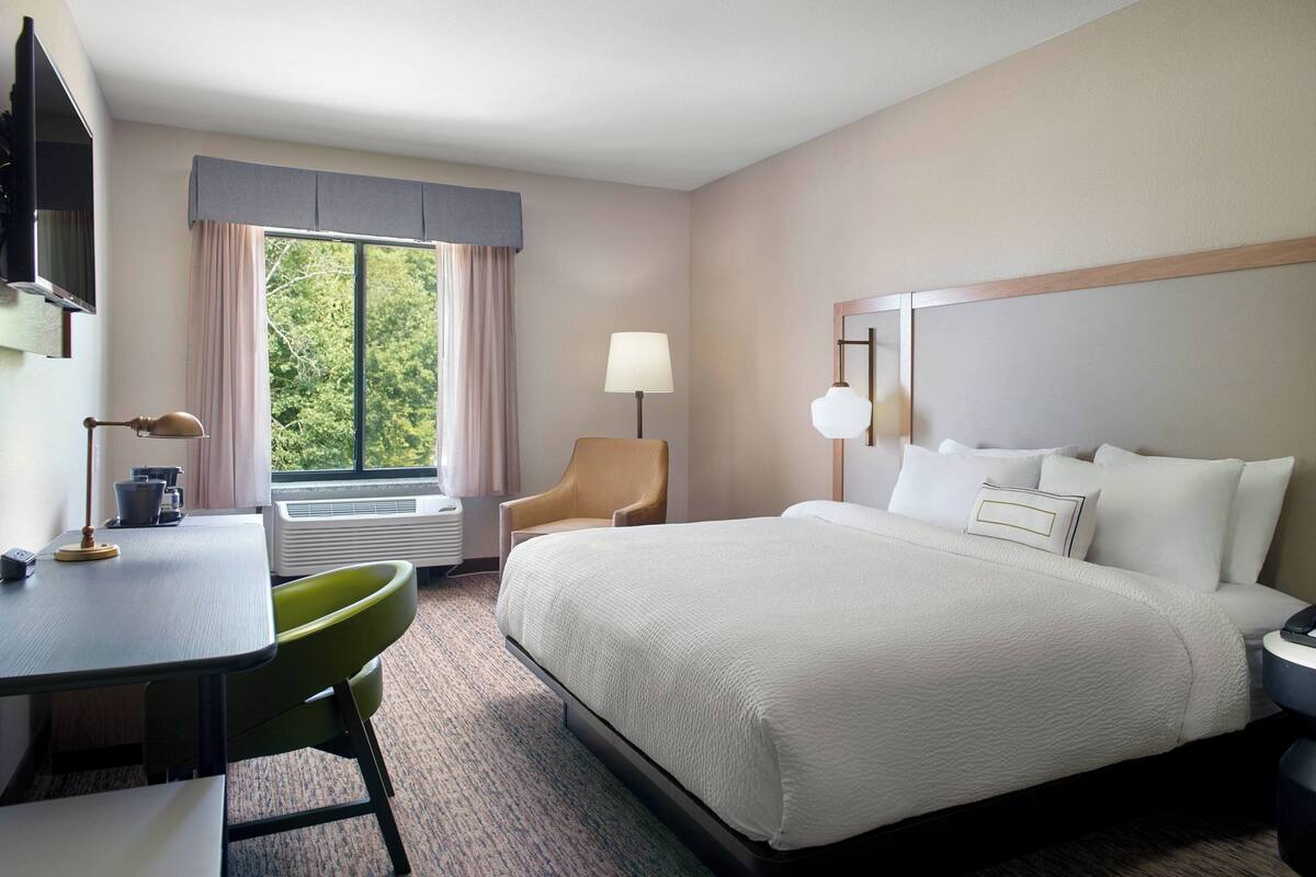 Une chambre d’hôtel avec un lit, un bureau, une chaise, une lampe et une fenêtre avec des rideaux.