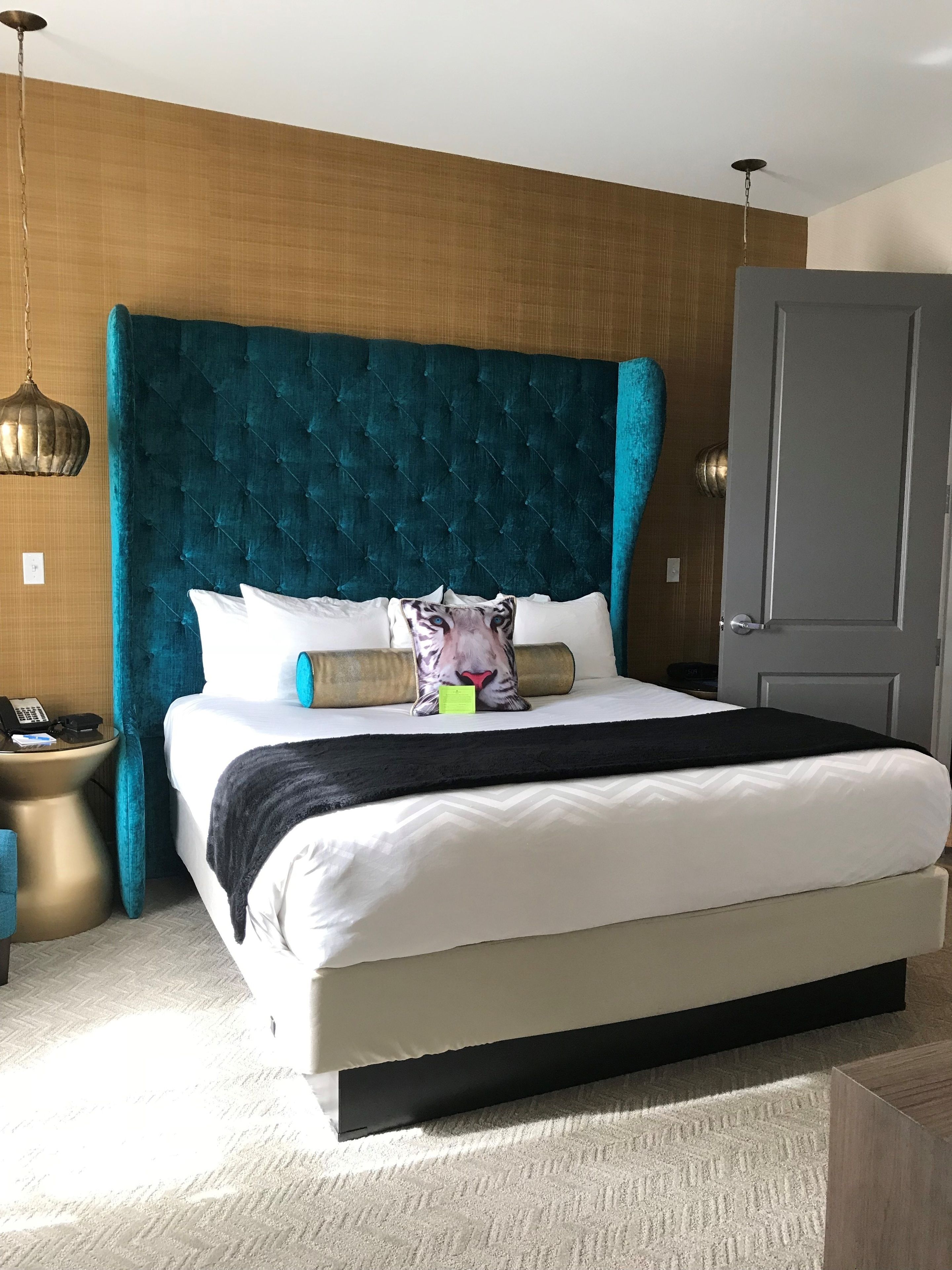 Una habitación de hotel moderna con una cama grande, un cabecero de madera, una mesita de noche con lámpara y una puerta.