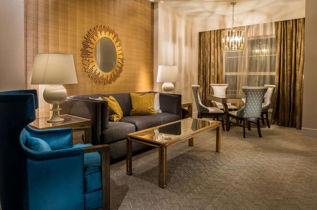 A hotel room with a sofa, armchairs, a coffee table, a dining table with chairs, a chandelier, and a large mirror on the wall.