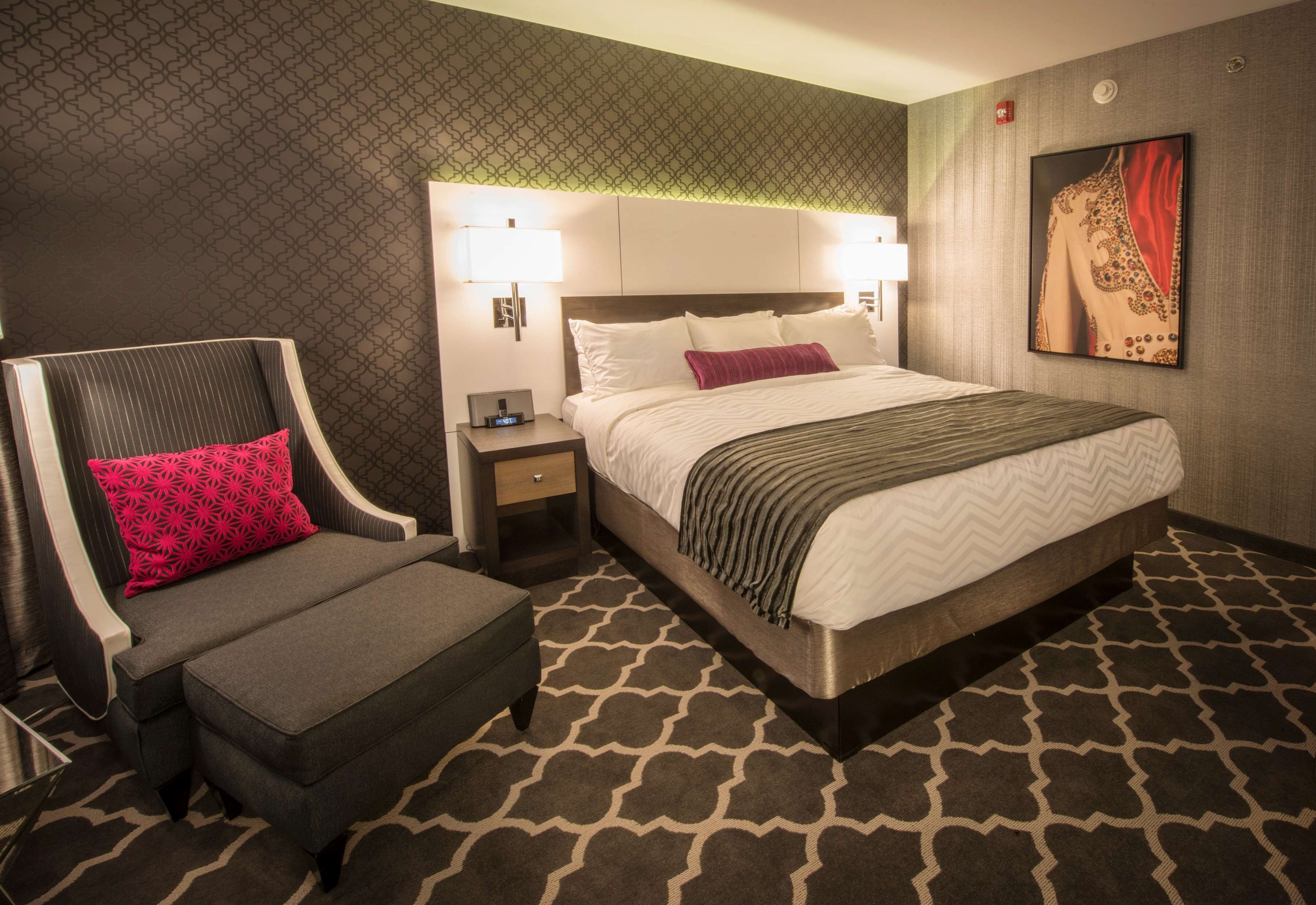 A hotel room with a large bed, a grey sofa with a red pillow, a nightstand with a lamp, and a patterned carpet.