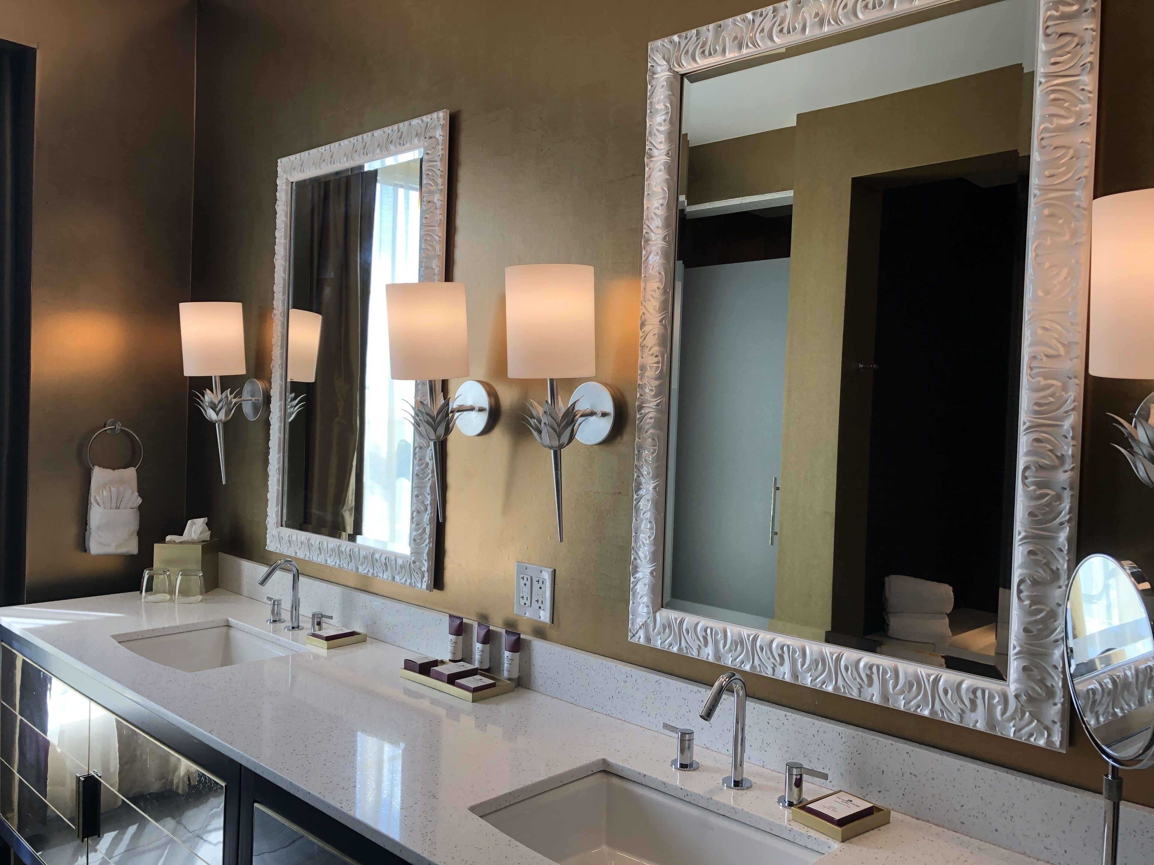 A hotel bathroom with two sinks, large mirrors, wall sconces, and a towel rack.