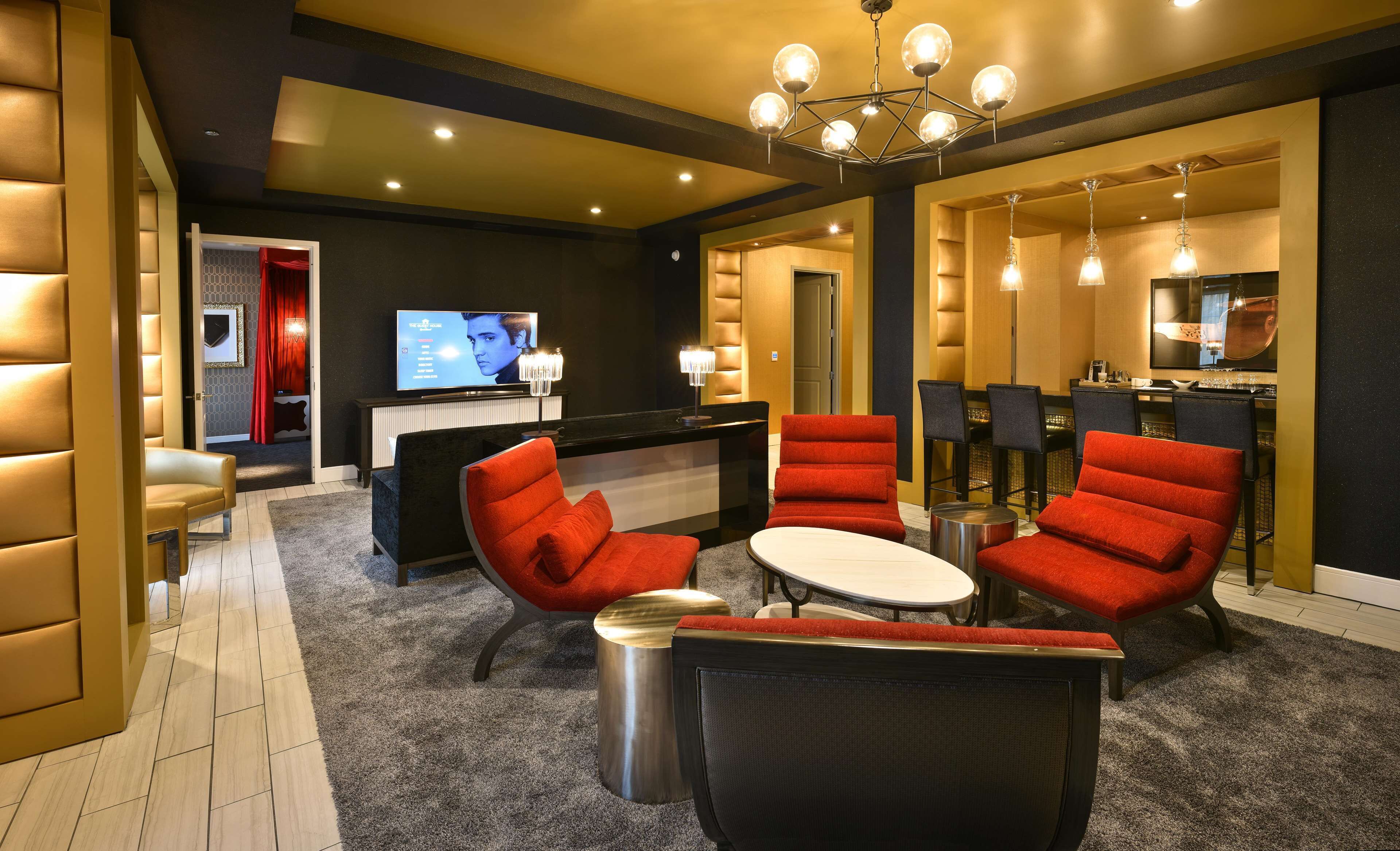 A hotel lobby with a reception desk, a television screen, red upholstered chairs, a round table, and a chandelier.