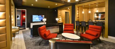 A hotel lobby with a reception desk, a television screen, red upholstered chairs, a round table, and a chandelier.