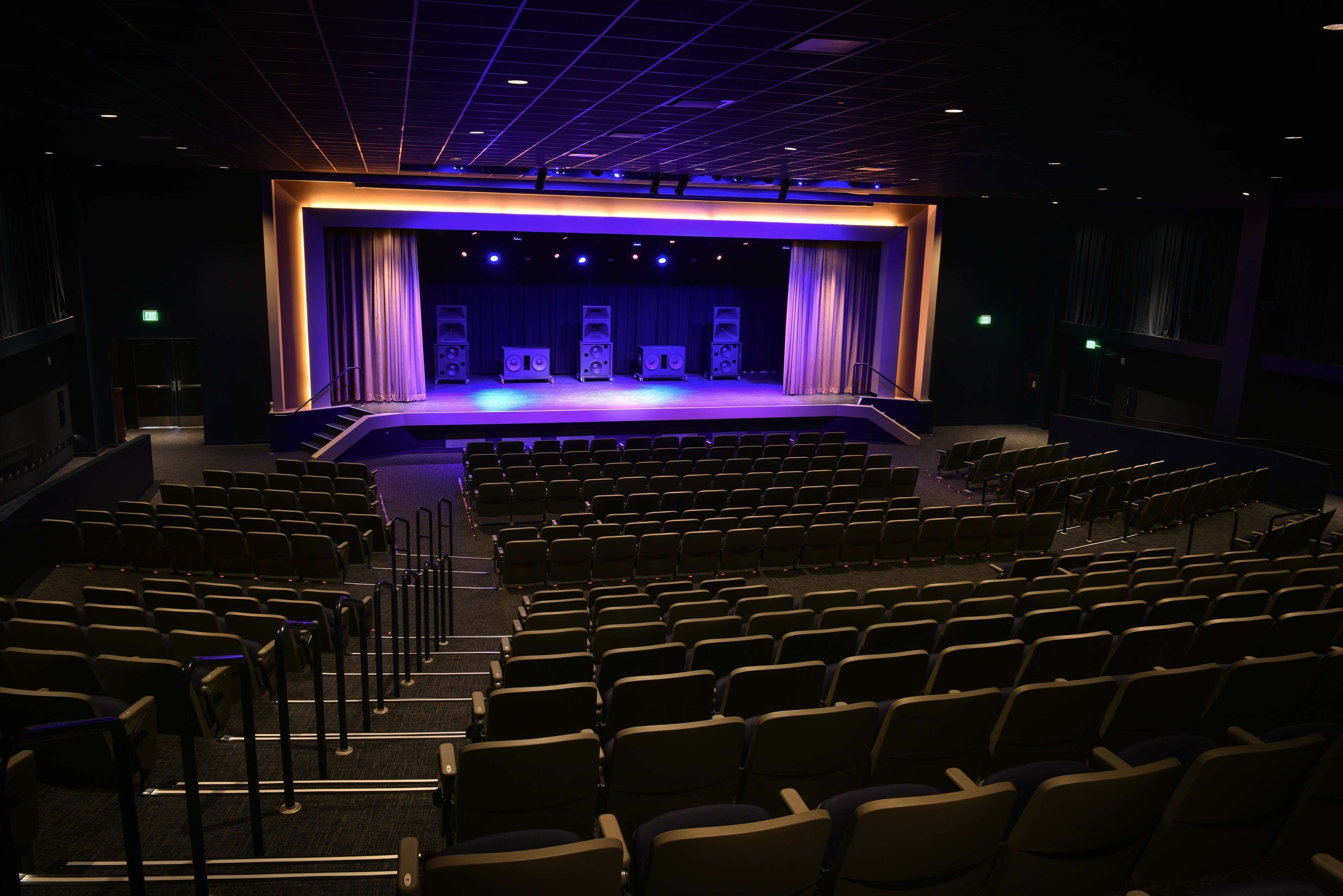An auditorium with rows of seats, a stage with curtains, and speakers.