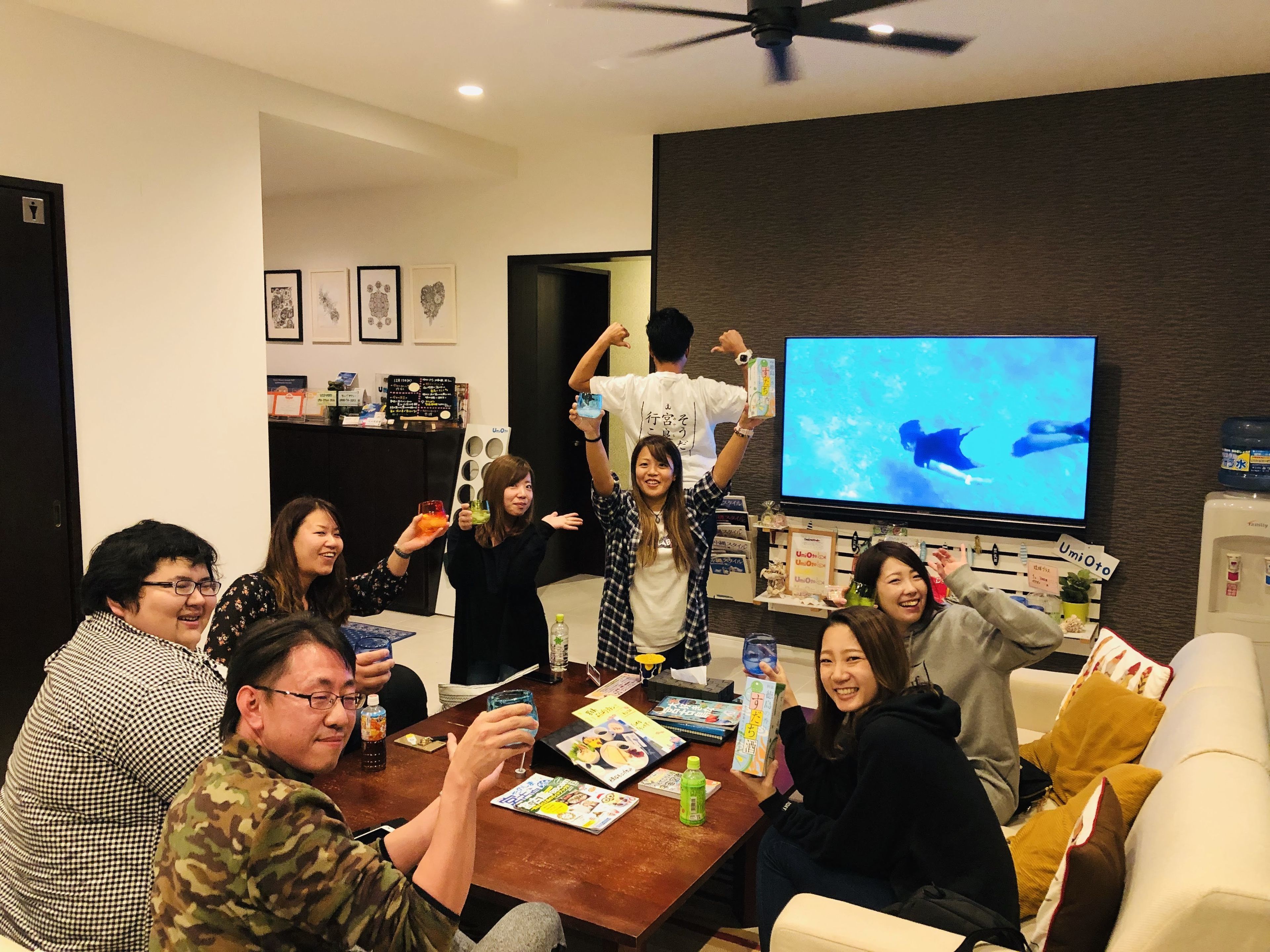 A group of people in a room with a television displaying a blue screen, a table with drinks and books, and a wall with framed pictures.