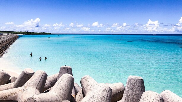 A beach with clear turquoise water, a rocky breakwater, and a distant shoreline.