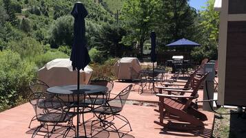 Patio esterno con tavoli e sedie in metallo, barbecue e ombrelloni.