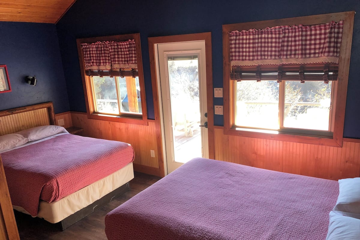 A room with two beds, wooden walls, and windows with checkered curtains.