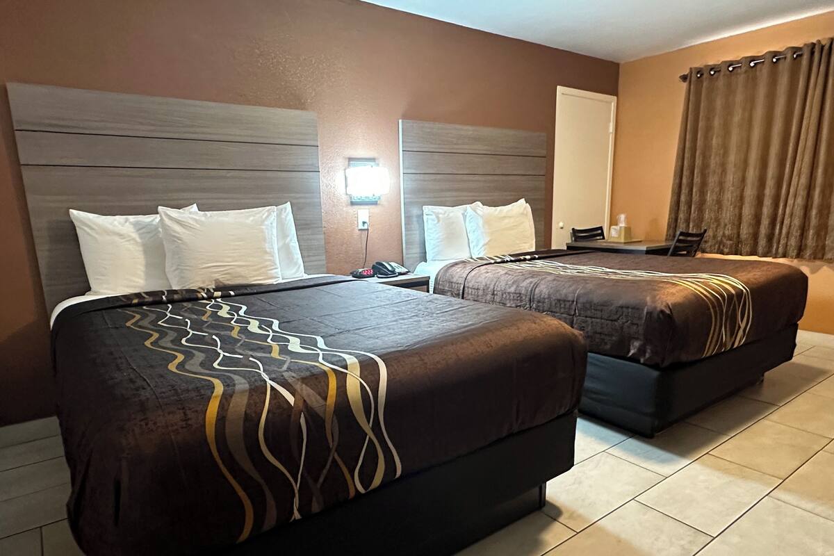 A hotel room with two beds, a nightstand, a lamp, and a window with curtains.