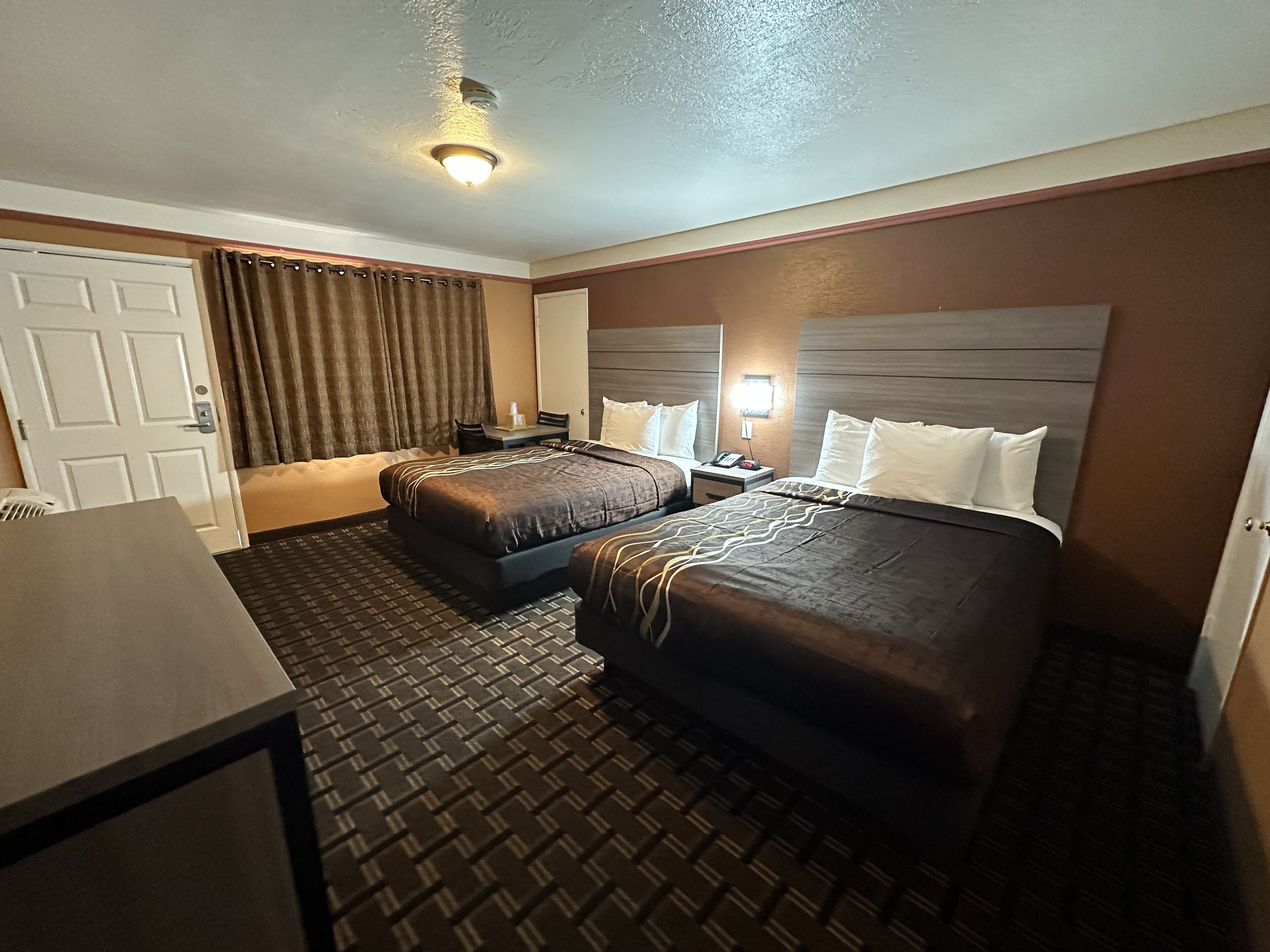 A hotel room with two beds, a desk, a chair, a nightstand, and a ceiling light.