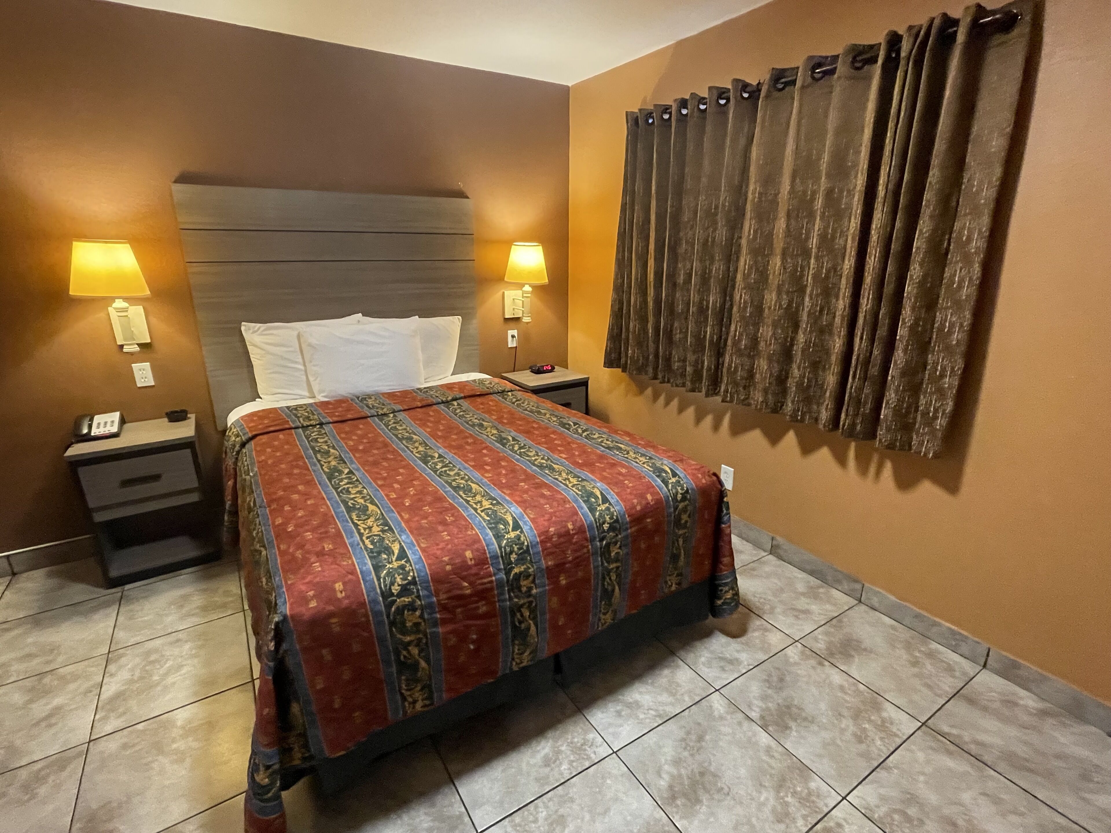 Una habitación de hotel con cama, dos mesitas de noche, lámpara, teléfono y cortina.