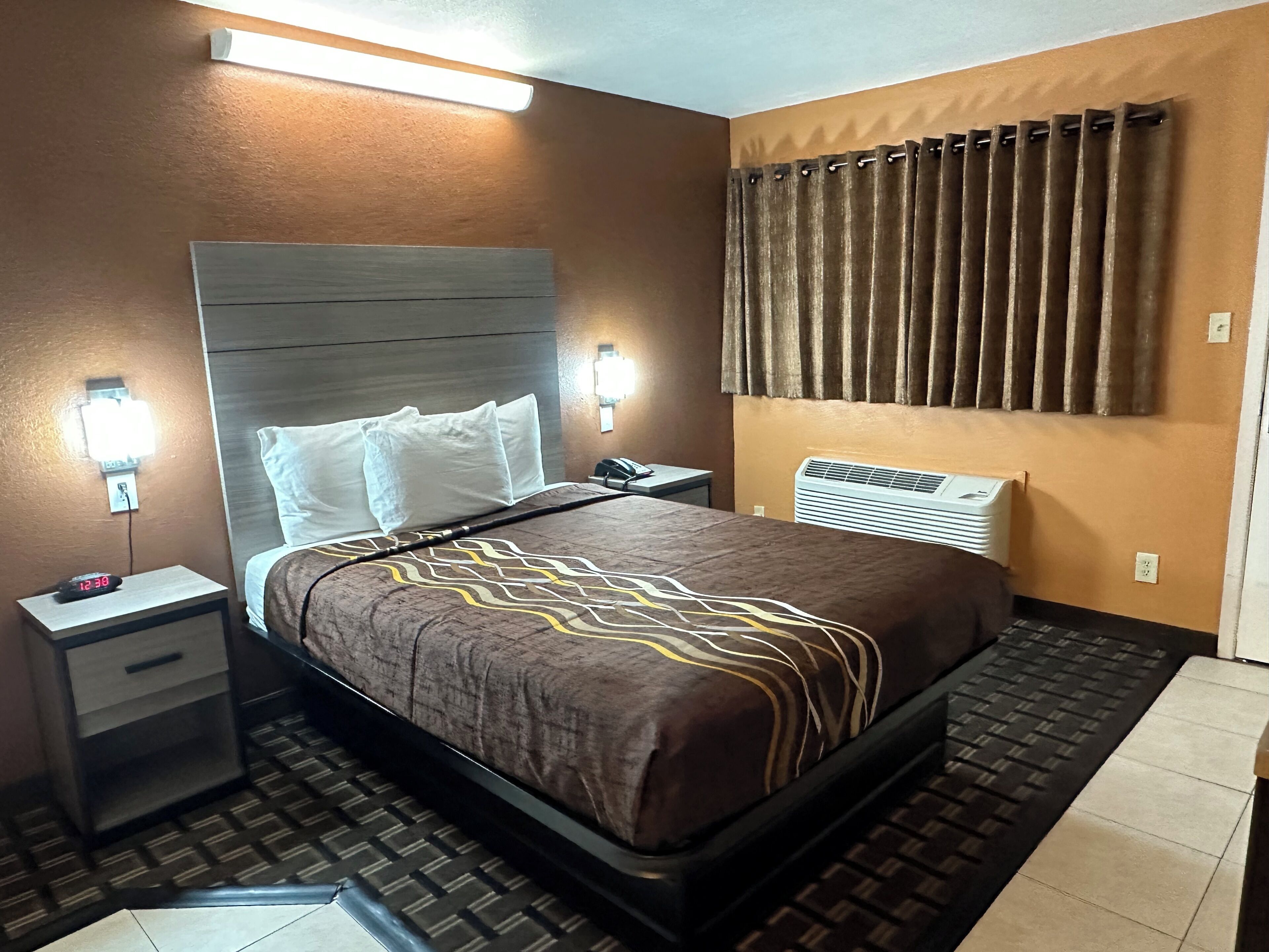A hotel room with a bed, bedside tables, a nightstand, a wall-mounted air conditioner, and a window with curtains.
