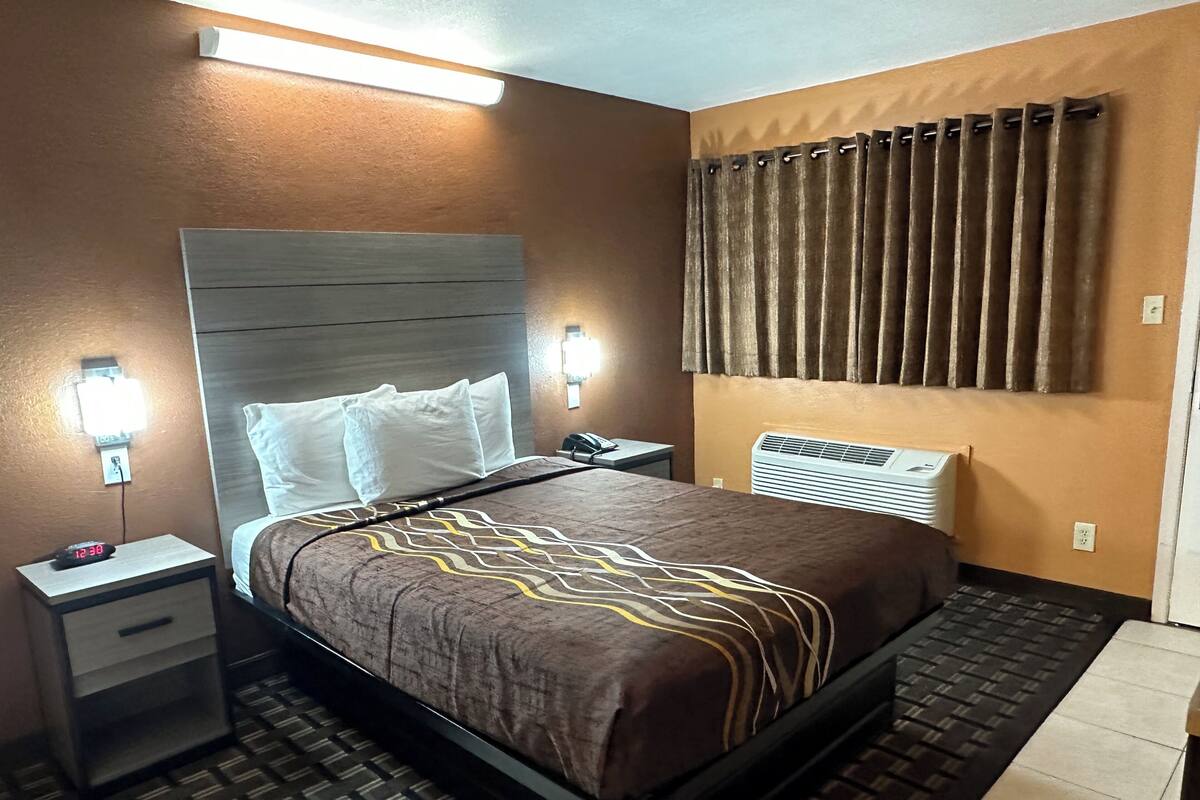 A hotel room with a bed, bedside tables, a nightstand, a wall-mounted air conditioner, and a window with curtains.
