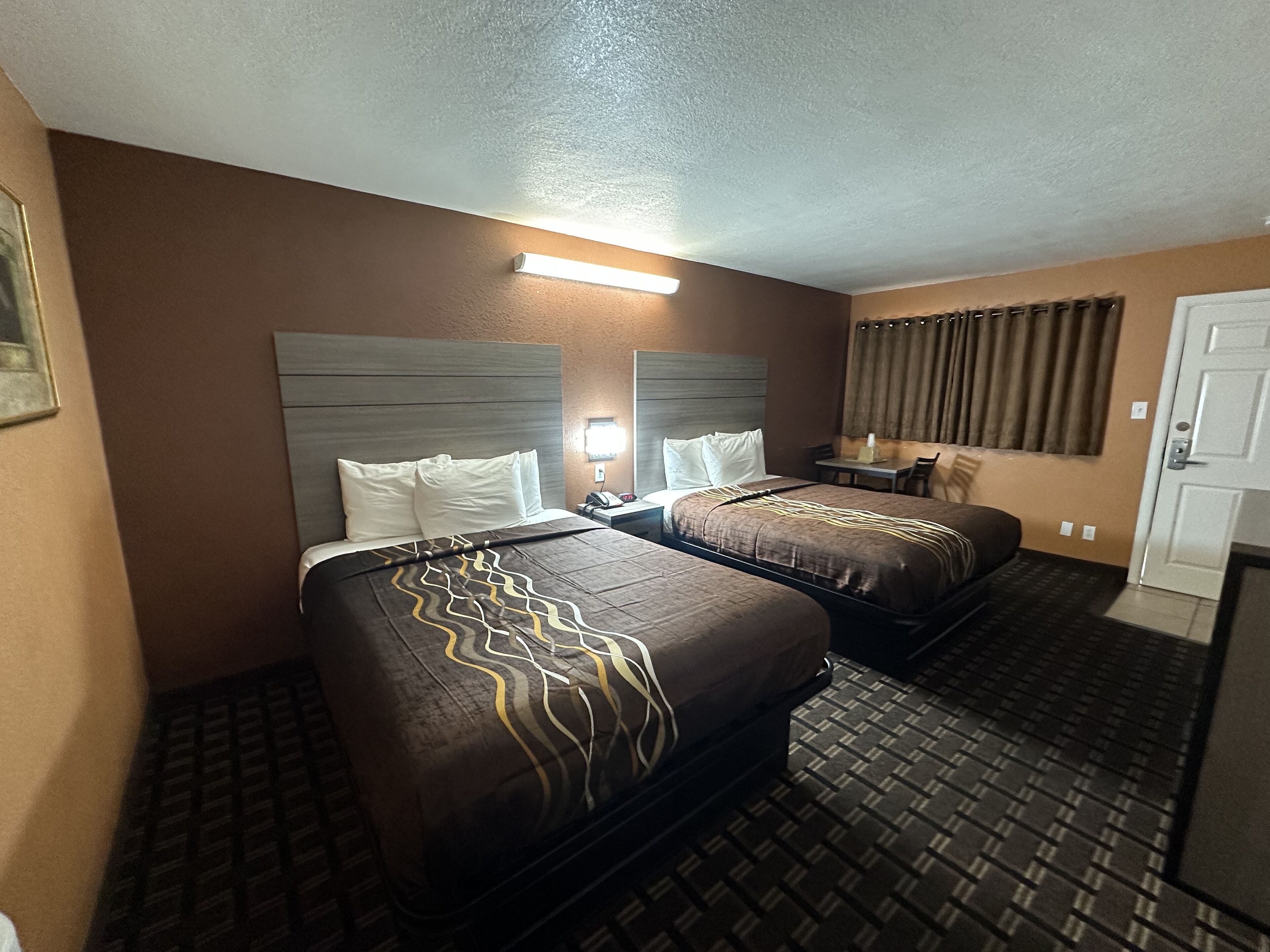 A hotel room with two beds, a nightstand, a lamp, a window with curtains, and a door.