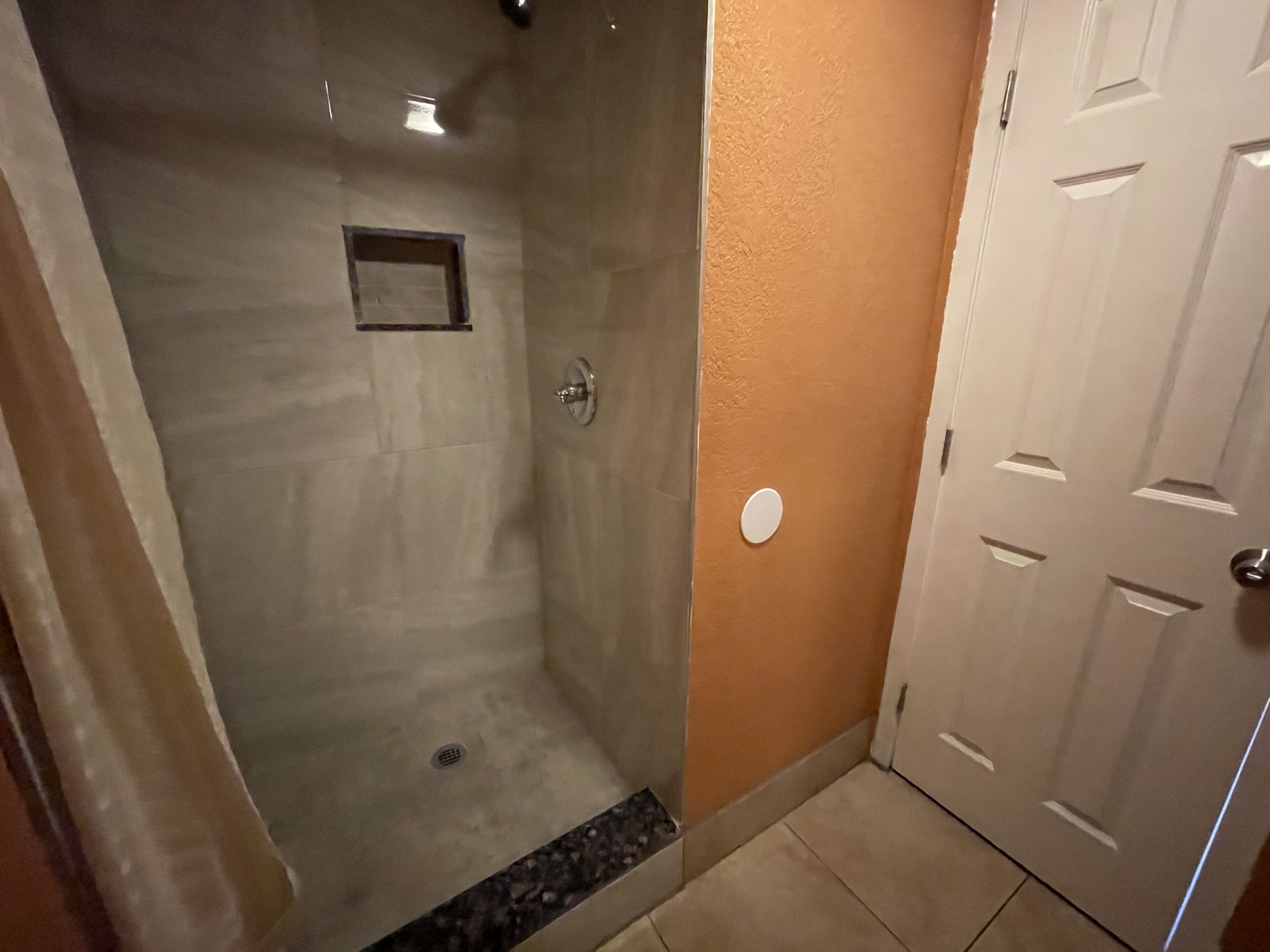 A shower with a glass door, a small square vent, and a white door to the right.