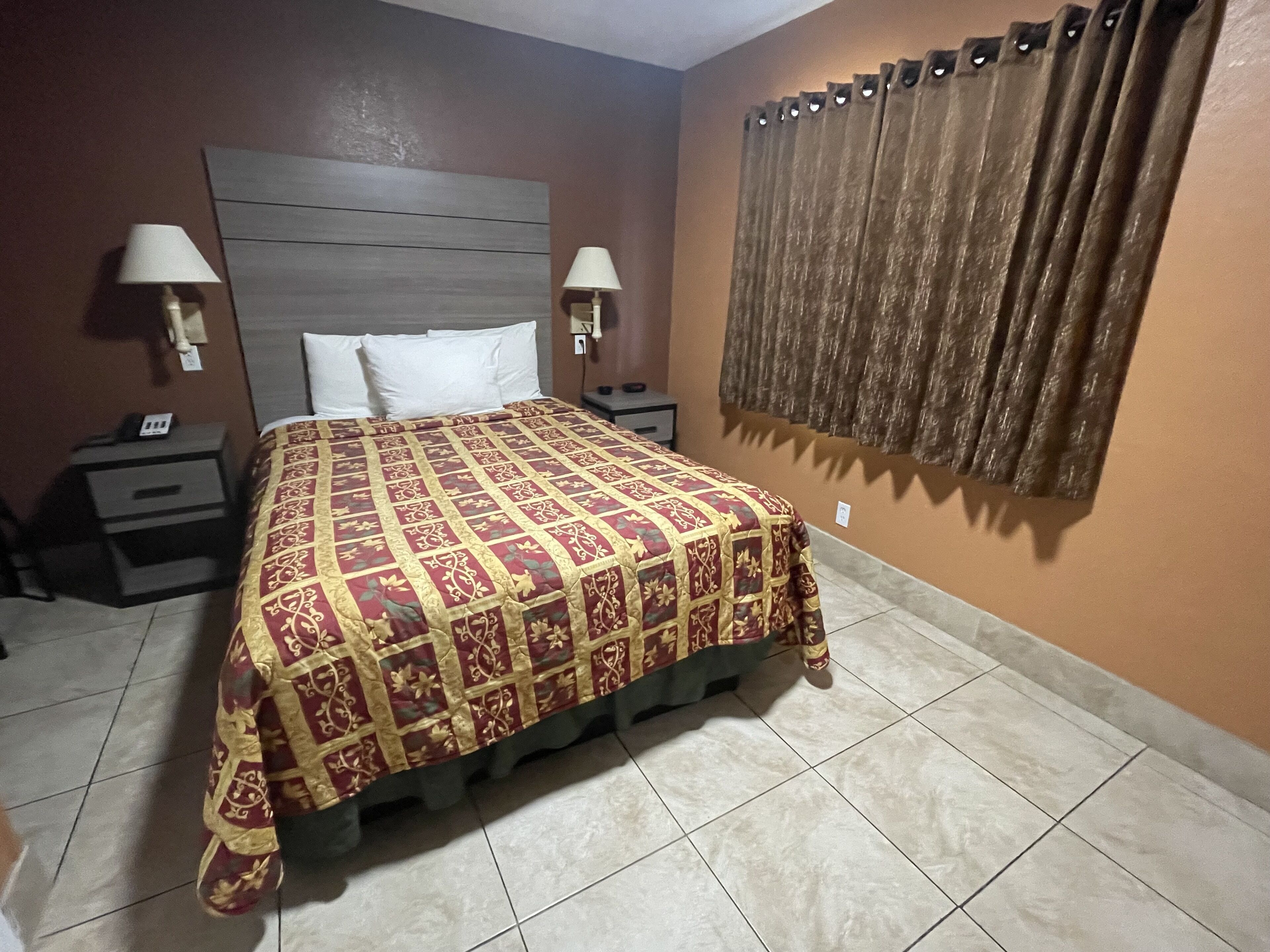 A hotel room with a large bed, two bedside tables with lamps, a headboard, a window with curtains, and a tiled floor.