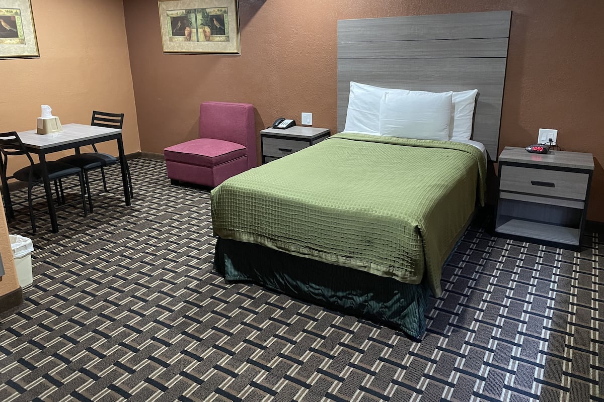 A hotel room with a bed, a desk with a chair, a small pink chair, a nightstand with a phone, and a wall-mounted picture.