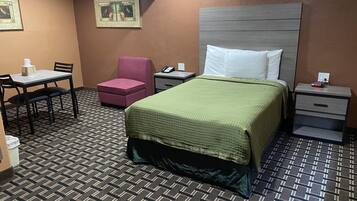 Ett hotellrum med en säng, ett skrivbord med en stol, en liten rosa stol, ett nattduksbord med en telefon och en tavla som hänger på väggen.